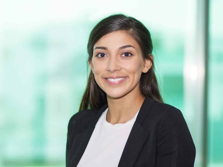 Una joven con el cabello castaño oscuro y largo, con un abrigo beige, está en una panadería sosteniendo una baguette y sonriendo a la cámara.